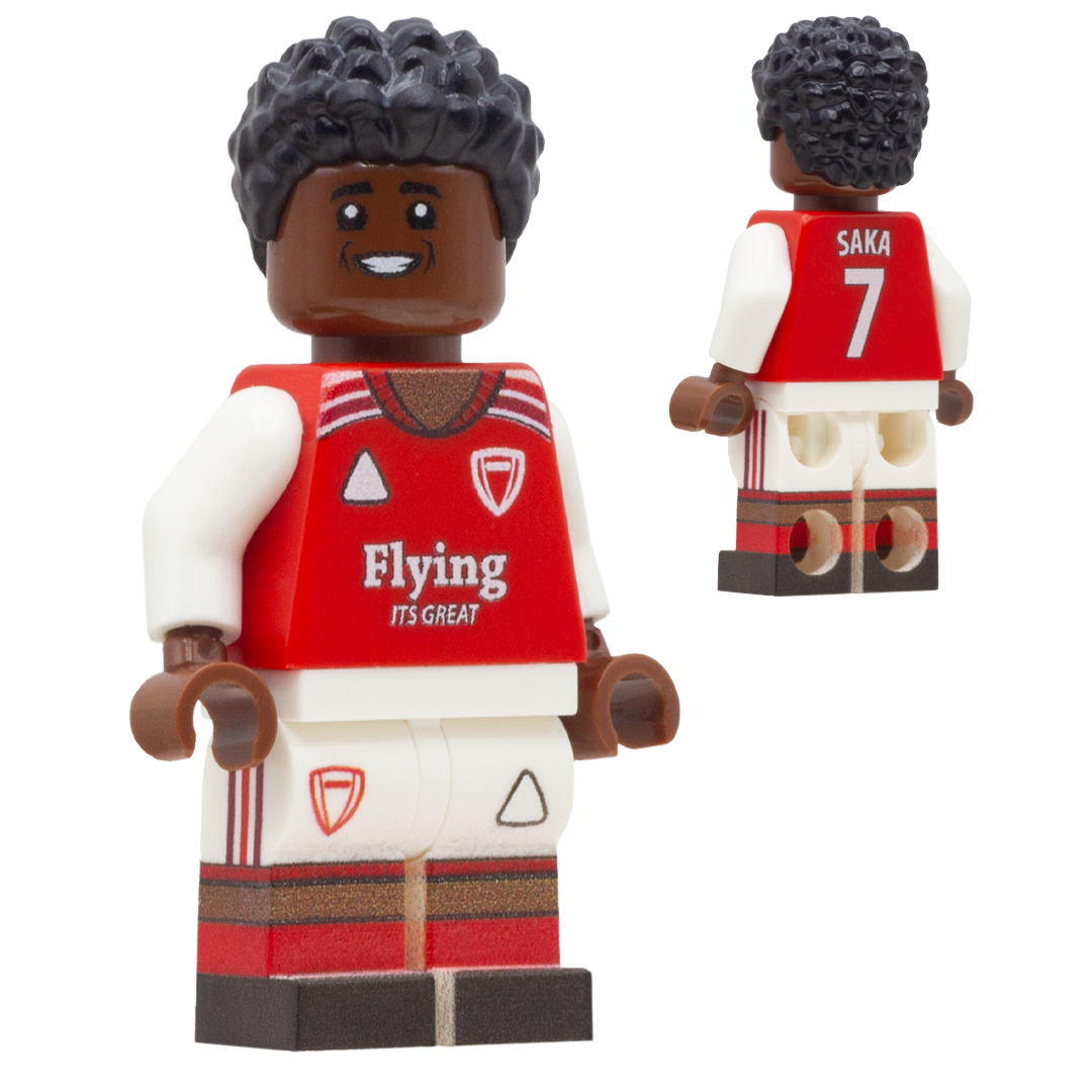 A toy soccer minifigure of Arsenal FC footballer Bukayo Saka. The figure has dark brown skin, black curly hair, a red and white Arsenal-style jersey with 'Flying' sponsor and a shield logo, white shorts, and red-and-white striped legs.