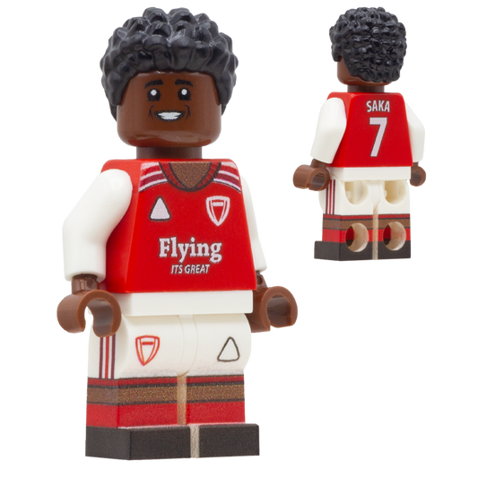 A toy soccer minifigure of Arsenal FC footballer Bukayo Saka. The figure has dark brown skin, black curly hair, a red and white Arsenal-style jersey with 'Flying' sponsor and a shield logo, white shorts, and red-and-white striped legs.