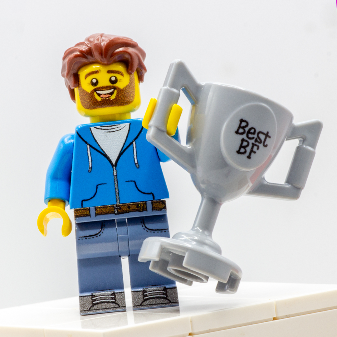 A custom LEGO minifigure with brown hair, a yellow face, a blue zip-up hoodie, and blue pants, holding a large gray trophy that says 'Best BF' against a white background.