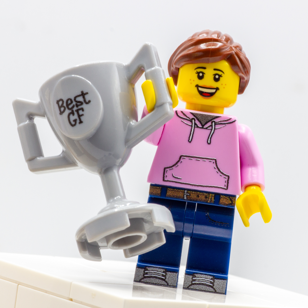 A custom LEGO minifigure with brown hair, a pink hoodie, and blue pants holding a large gray trophy that says 'Best GF'.