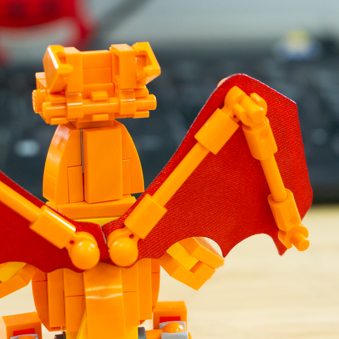 A close-up of the back of a toy dragon built from orange and red building blocks, with wings raised, set against a blurred background.