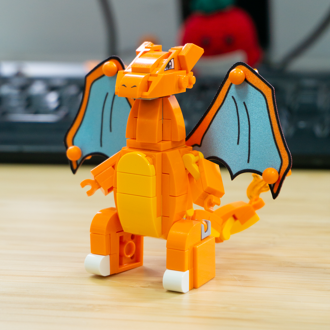A custom LEGO Charizard figure built from orange and yellow building blocks, with light blue wings outlined in black, standing on a light wood surface with a blurred keyboard in the background.
