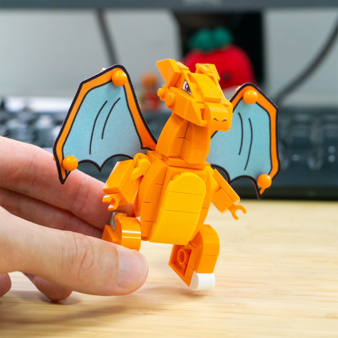 A hand holds a small orange dragon toy made of interlocking building blocks, with light blue wings outlined in black, on a wooden tabletop with a blurred background.