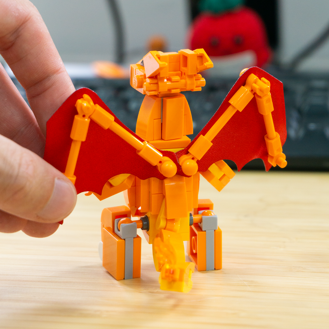 A hand is holding an orange and red dragon toy built from interlocking blocks on a light wooden tabletop, with a blurred background showing a desk and a red plush toy. The dragon is shown from the back. 