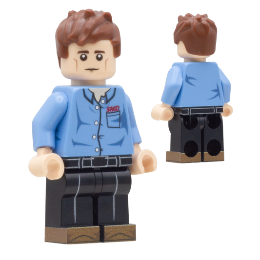 A toy minifigure shown from the front and back on a plain white background. The figure has brown hair, a beige face, a light blue button-up shirt with red 'SMD' lettering on the chest, black pants, and tan shoes.