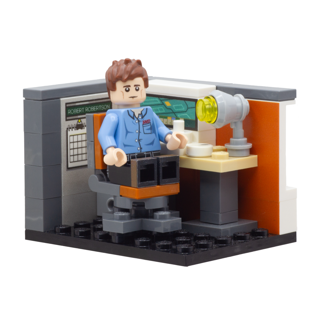 A small brick-built scene featuring a minifigure seated at a desk with a lamp, set against a grey and orange wall backdrop on a black LEGO baseplate.