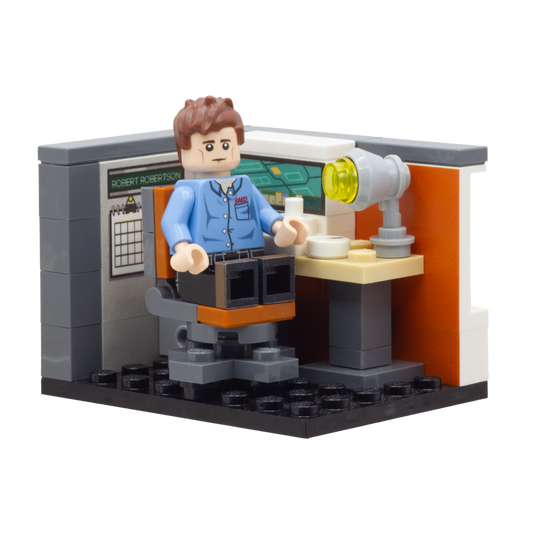 A small brick-built scene featuring a minifigure seated at a desk with a lamp, set against a grey and orange wall backdrop on a black LEGO baseplate.