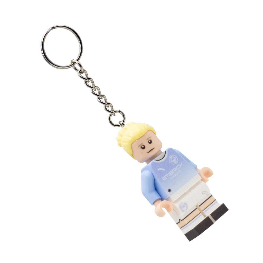 A soccer player keychain featuring a small plastic minifigure with blonde hair, wearing a light blue and white Manchester City jersey, attached to a silver metal split ring on a white background.