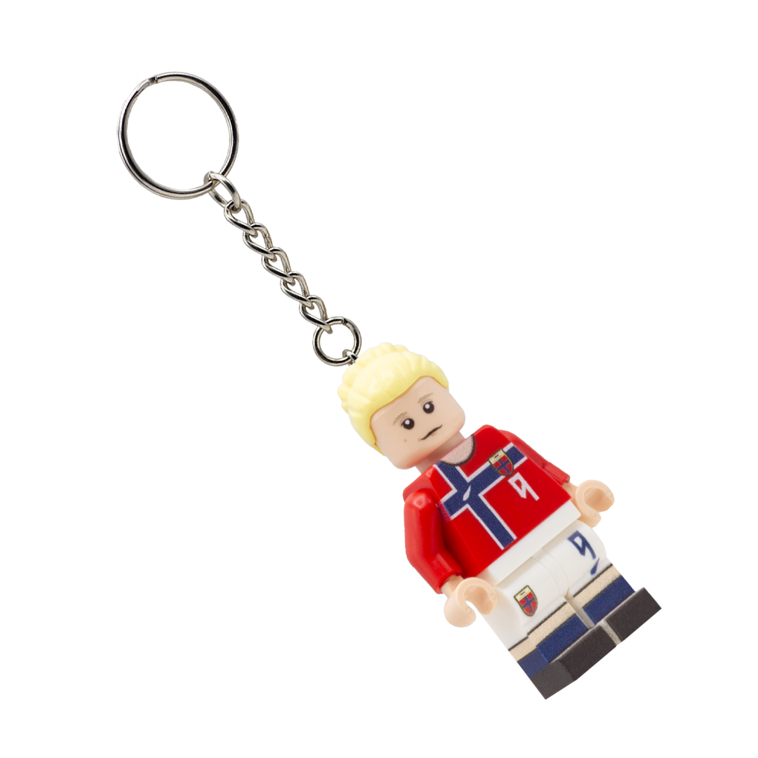 A miniature soccer player keychain charm with blond hair, wearing a red jersey with a blue and white cross, white shorts, and black and blue striped socks, attached to a silver metal key ring and chain on a white background.
