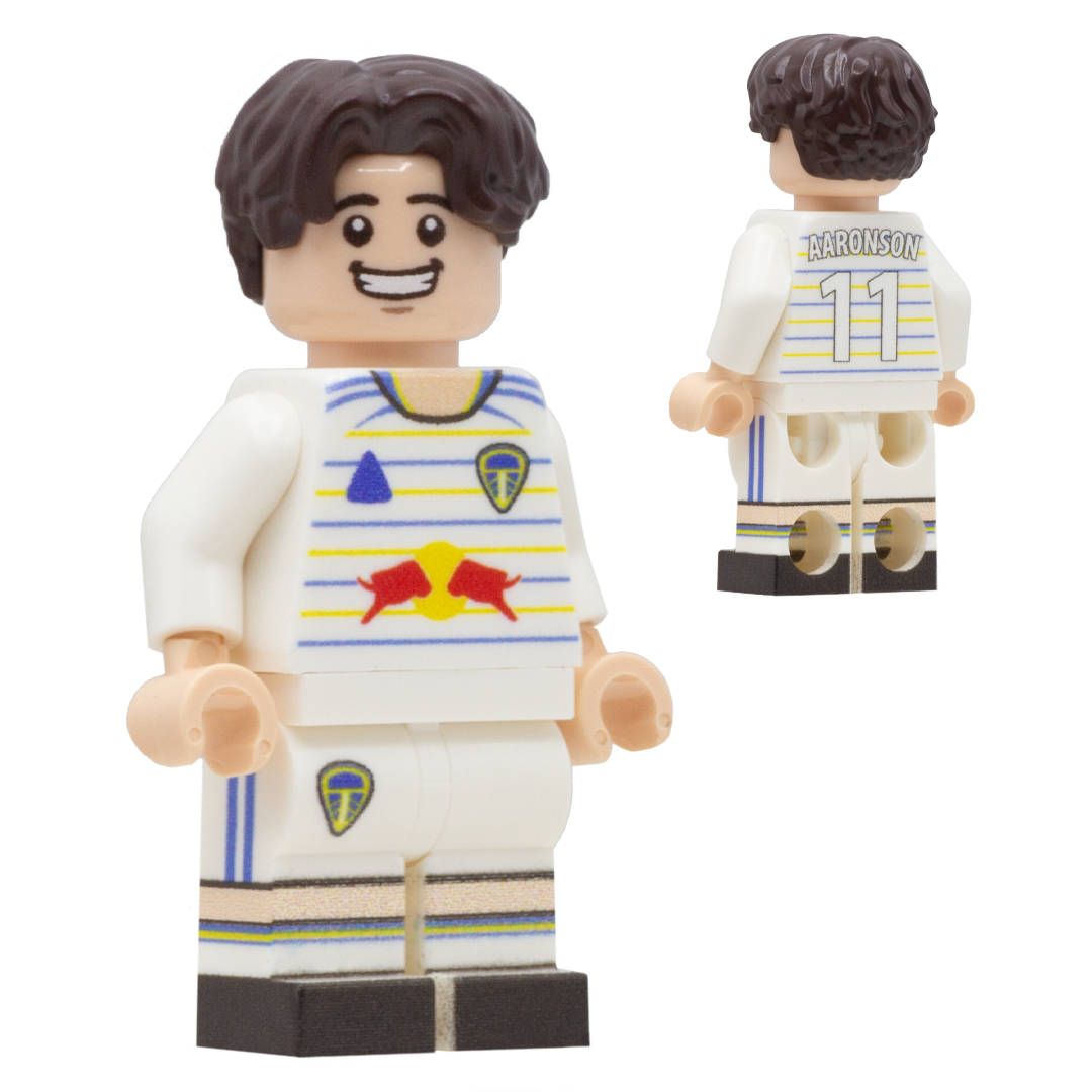 A LEGO-style soccer minifigure of Leeds United FC footballer, Brenden Aaronson. The figure has dark brown hair, a light complexion, a white jersey with blue and yellow stripes, a red bull logo sponsor.
