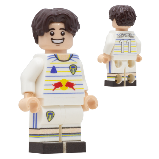 A LEGO-style soccer minifigure of Leeds United FC footballer, Brenden Aaronson. The figure has dark brown hair, a light complexion, a white jersey with blue and yellow stripes, a red bull logo sponsor.