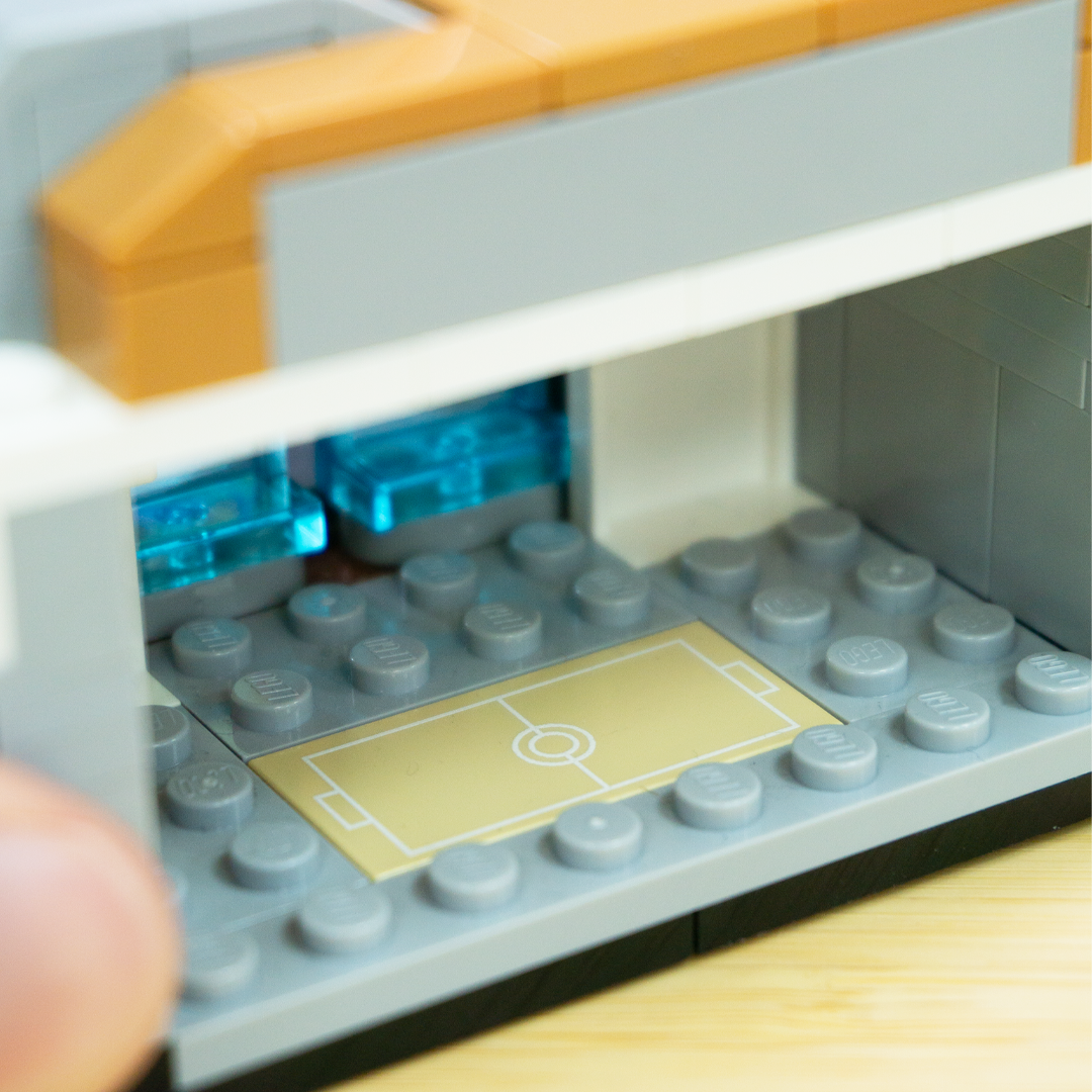 Close-up of a LEGO-style building set showing a gray baseplate with a small light tan Pokemon training area, translucent blue pieces, and orange and white upper structure. A finger is visible at the lower left edge.