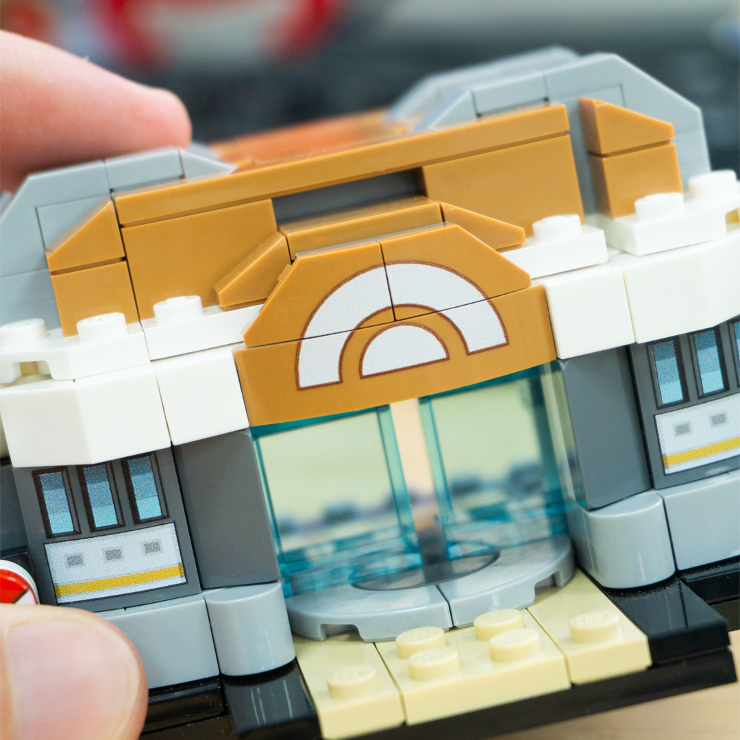 Close-up of a toy building-block structure being assembled by hands, showing tan, white, gray, and light blue pieces with a small light tan base.
