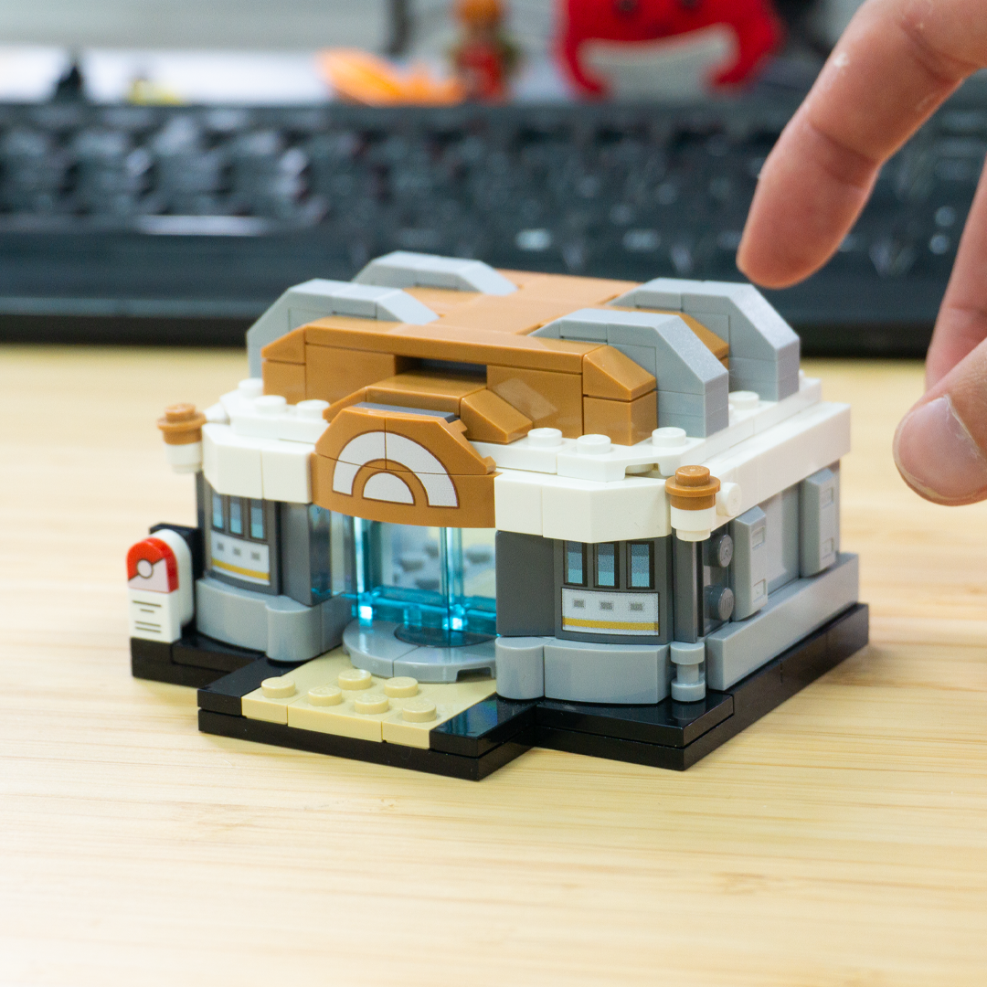 A small brick-built building model with a tan and white facade, gray roof details, and a clear blue glass entrance, shown on a light wood desk with a finger pointing at it. A blurred keyboard and red object appear in the background.