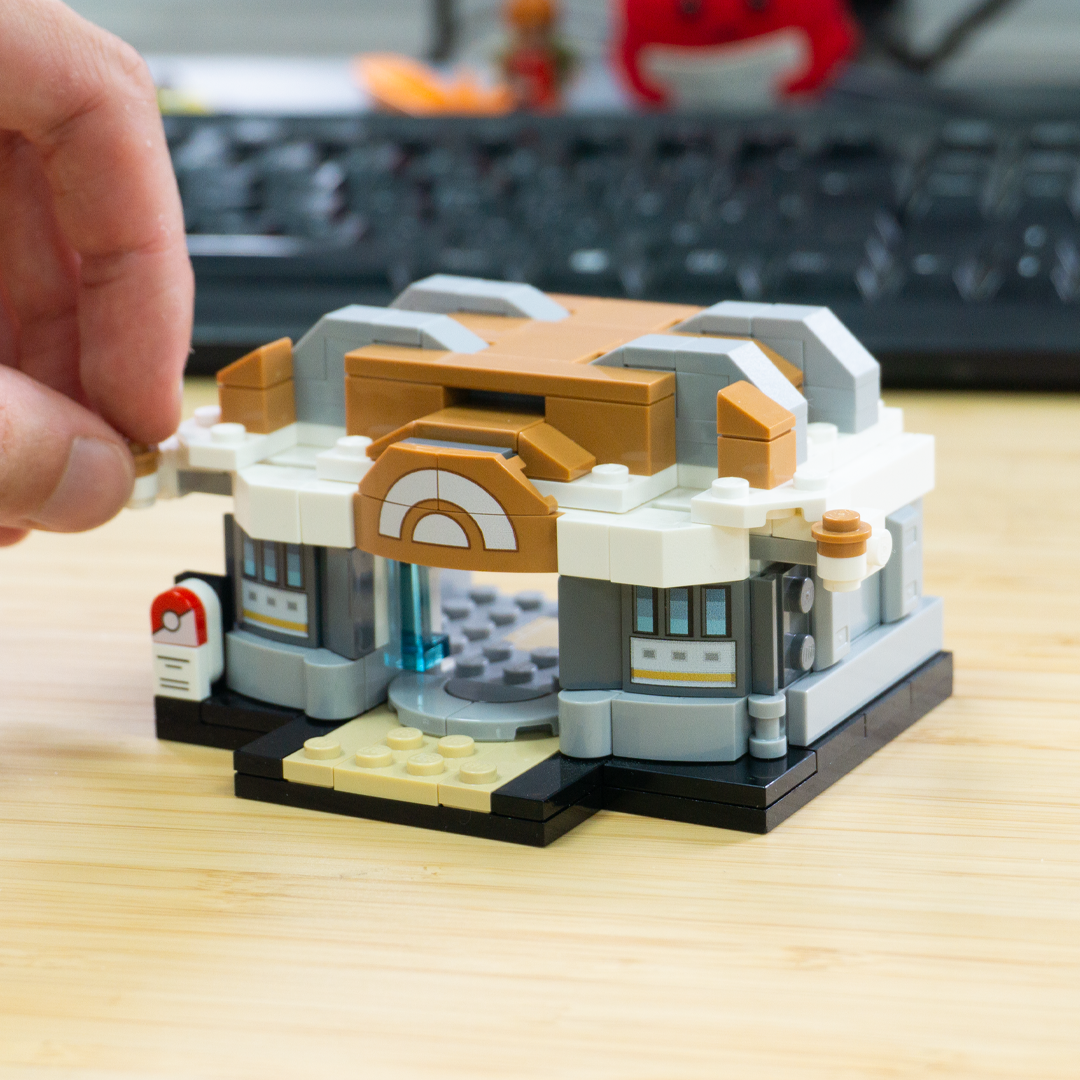 A hand is building a small custom LEGO Pokemon training gym on a light wood desk, with a blurred keyboard and desk background behind it.