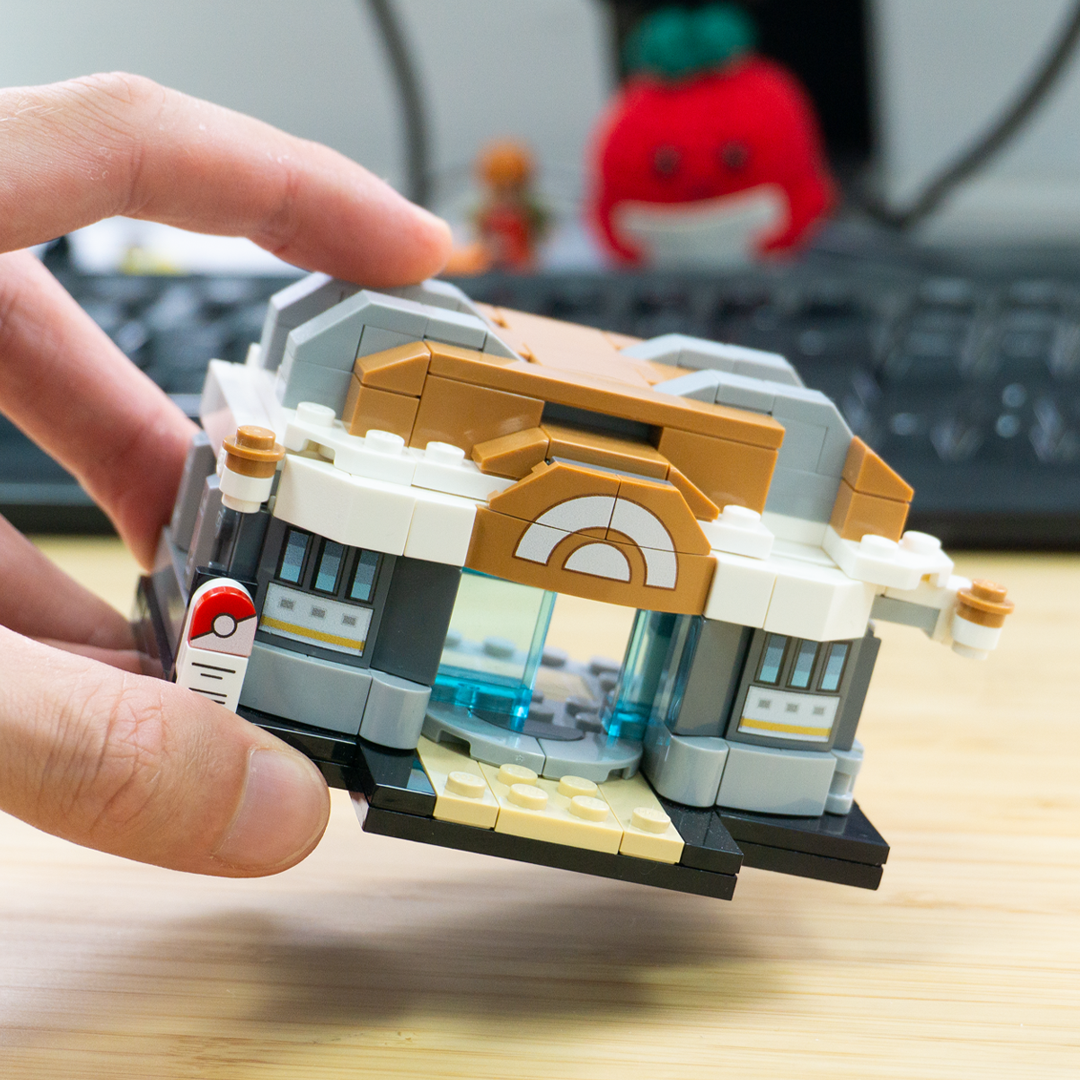 A hand holds a small custom-built toy building set resembling a Pokéball-themed structure, with a blurred keyboard and red plush in the background.