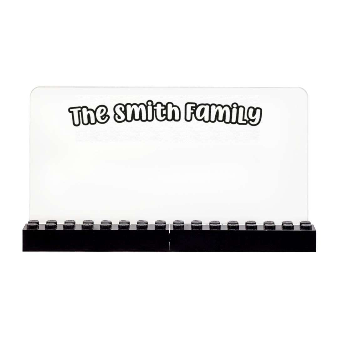 A personalized clear acrylic family name sign with black LEGO bricks along the bottom, featuring the text 'The smith Family' in a white font with a black outline.