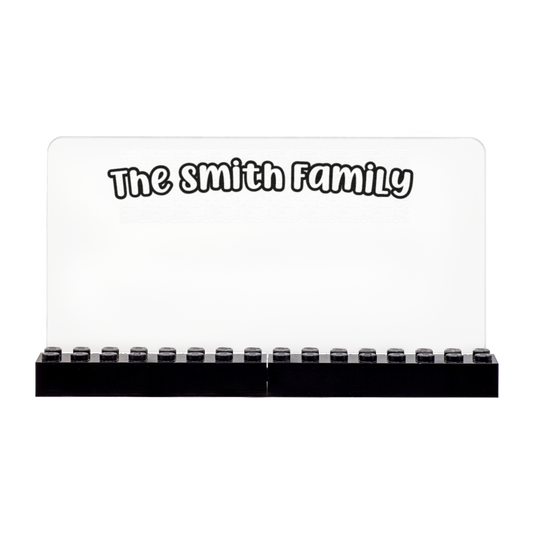 A personalized clear acrylic family name sign with black LEGO bricks along the bottom, featuring the text 'The smith Family' in a white font with a black outline.