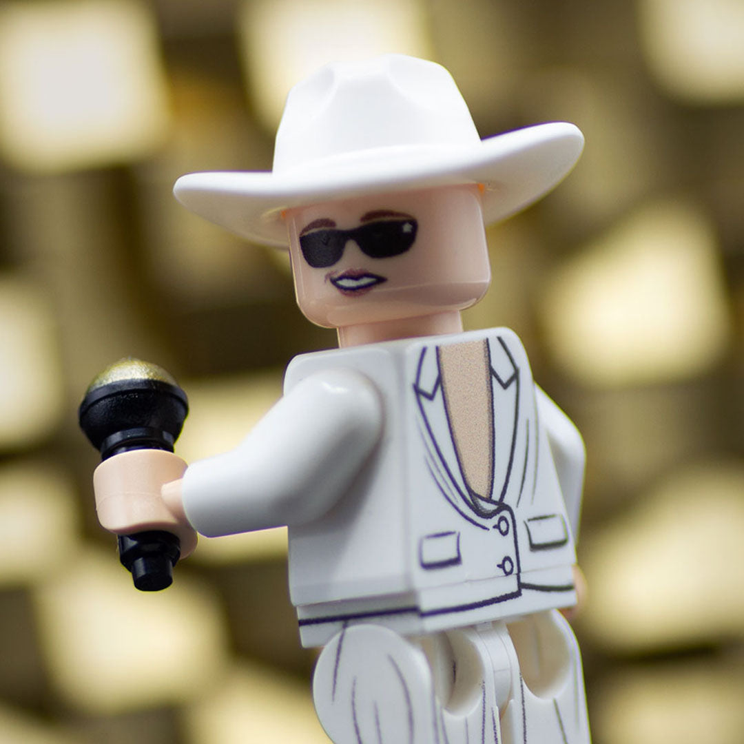 A close-up of a toy minifigure (based on Celine Dion) wearing a white cowboy hat, black sunglasses, and a white backwards suit with black line details, holding a black and gold microphone against a blurred warm background.