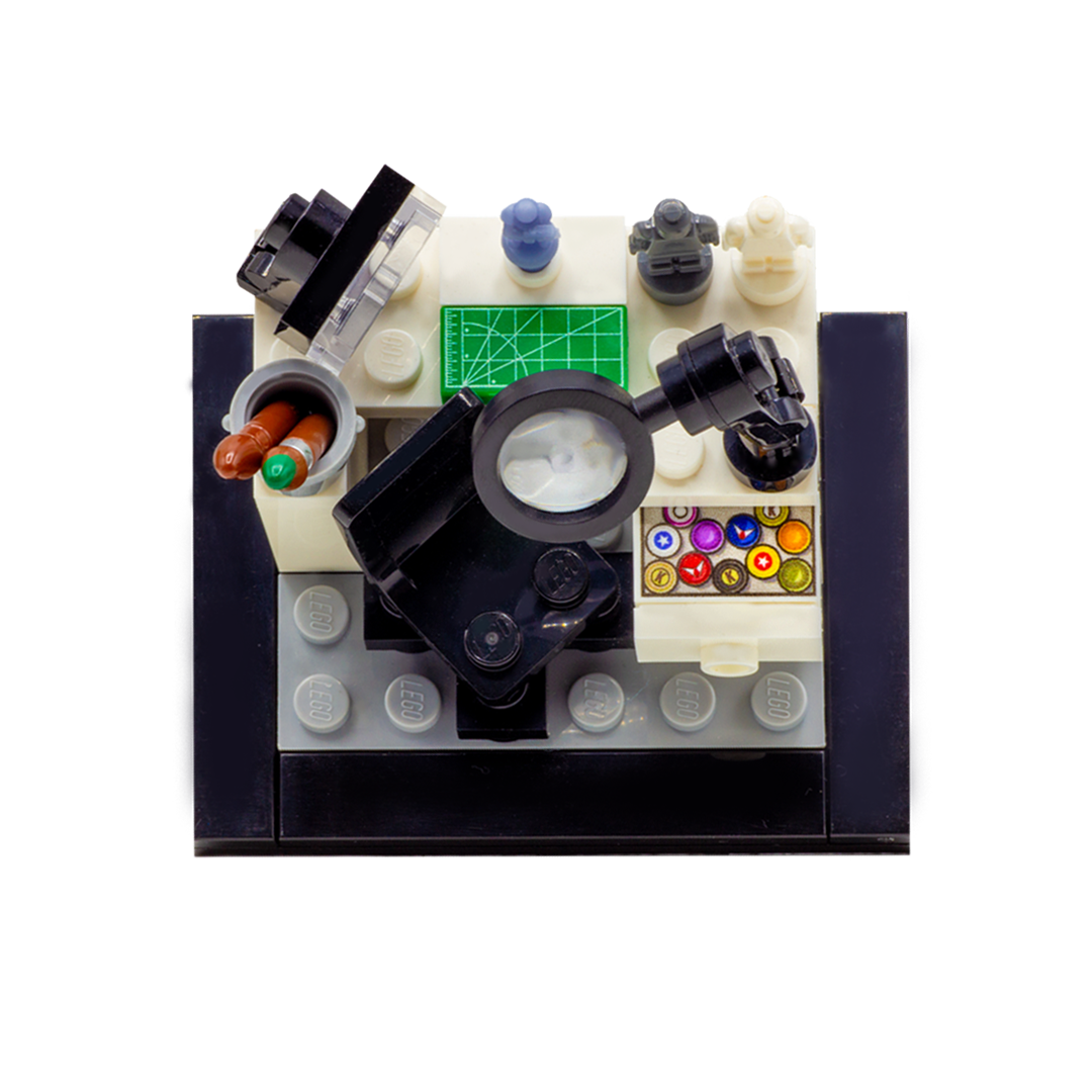A LEGO-style miniature artist's workbench set on a dark grey and black base, featuring a white desk, black magnifying glass, lamp, paintbrushes, miniature figures, accessories and custom-printed tiles. Top view