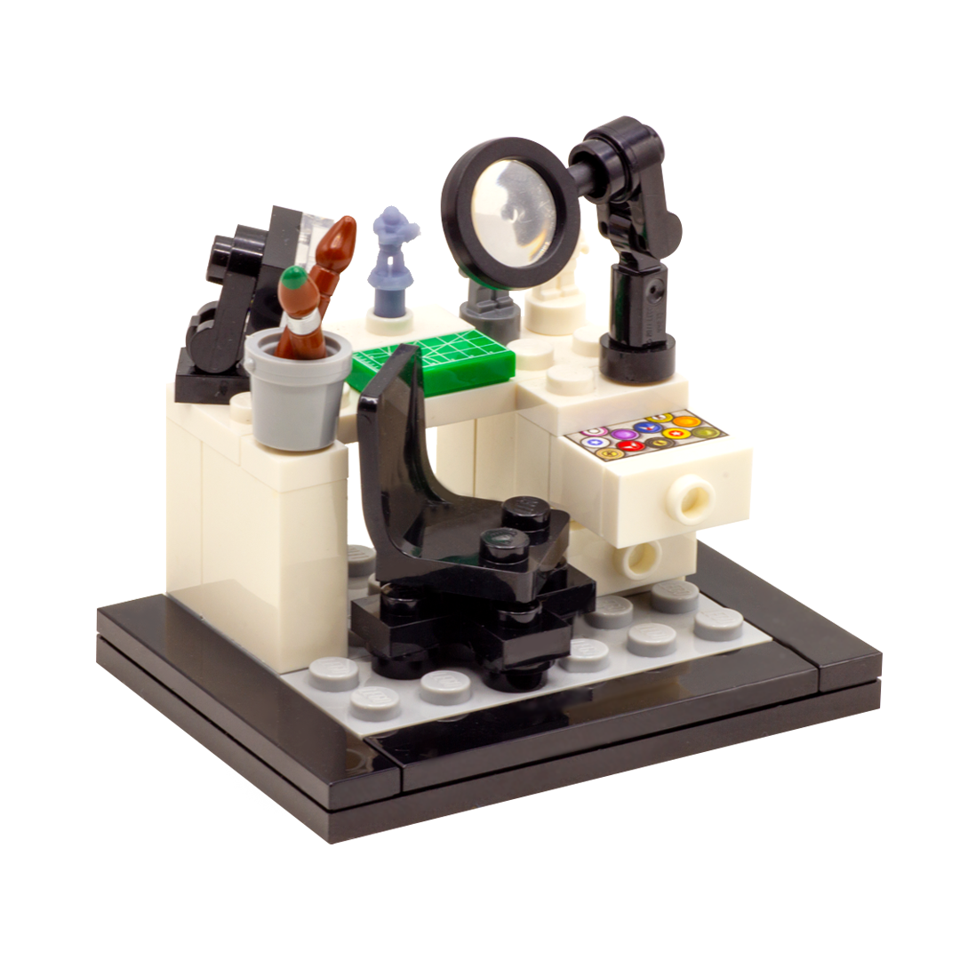 A LEGO-style miniature artist's workbench set on a dark grey and black base, featuring a white desk, black magnifying glass, lamp, paintbrushes, miniature figures, accessories and custom-printed tiles.
