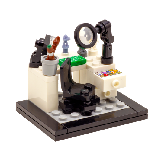 A LEGO-style miniature artist's workbench set on a dark grey and black base, featuring a white desk, black magnifying glass, lamp, paintbrushes, miniature figures, accessories and custom-printed tiles.