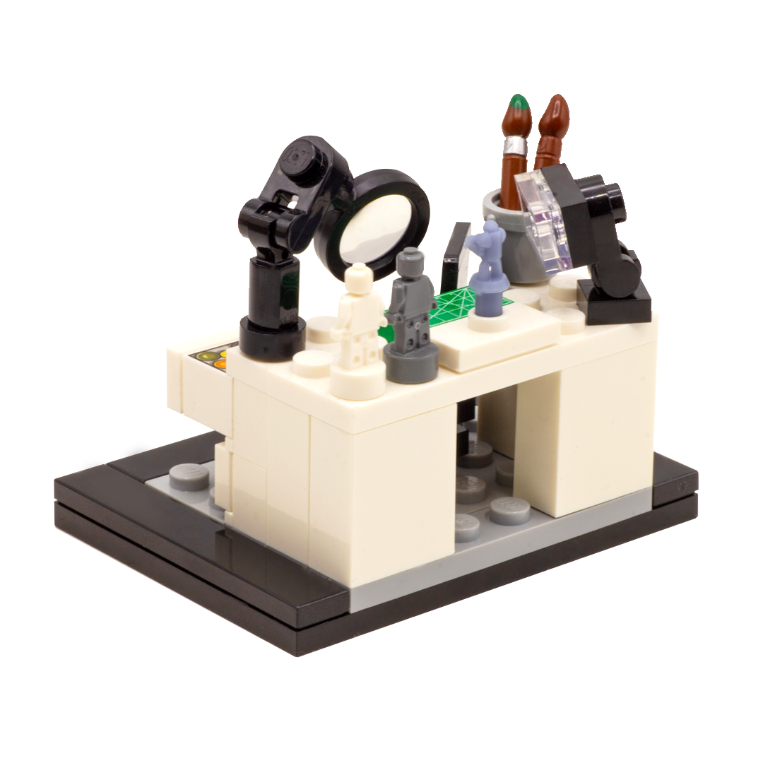 A LEGO-style miniature artist's workbench set on a dark grey and black base, featuring a white desk, black magnifying glass, lamp, paintbrushes, miniature figures, accessories and custom-printed tiles. Rear view.