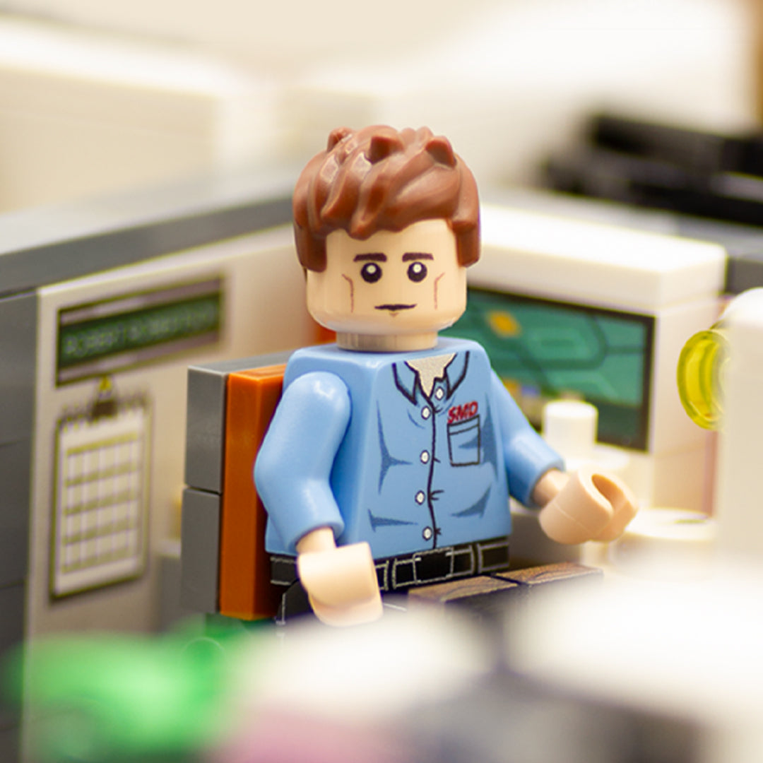A small brick-built scene featuring a minifigure seated at a desk with a lamp, set against a grey and orange wall backdrop on a black LEGO baseplate.