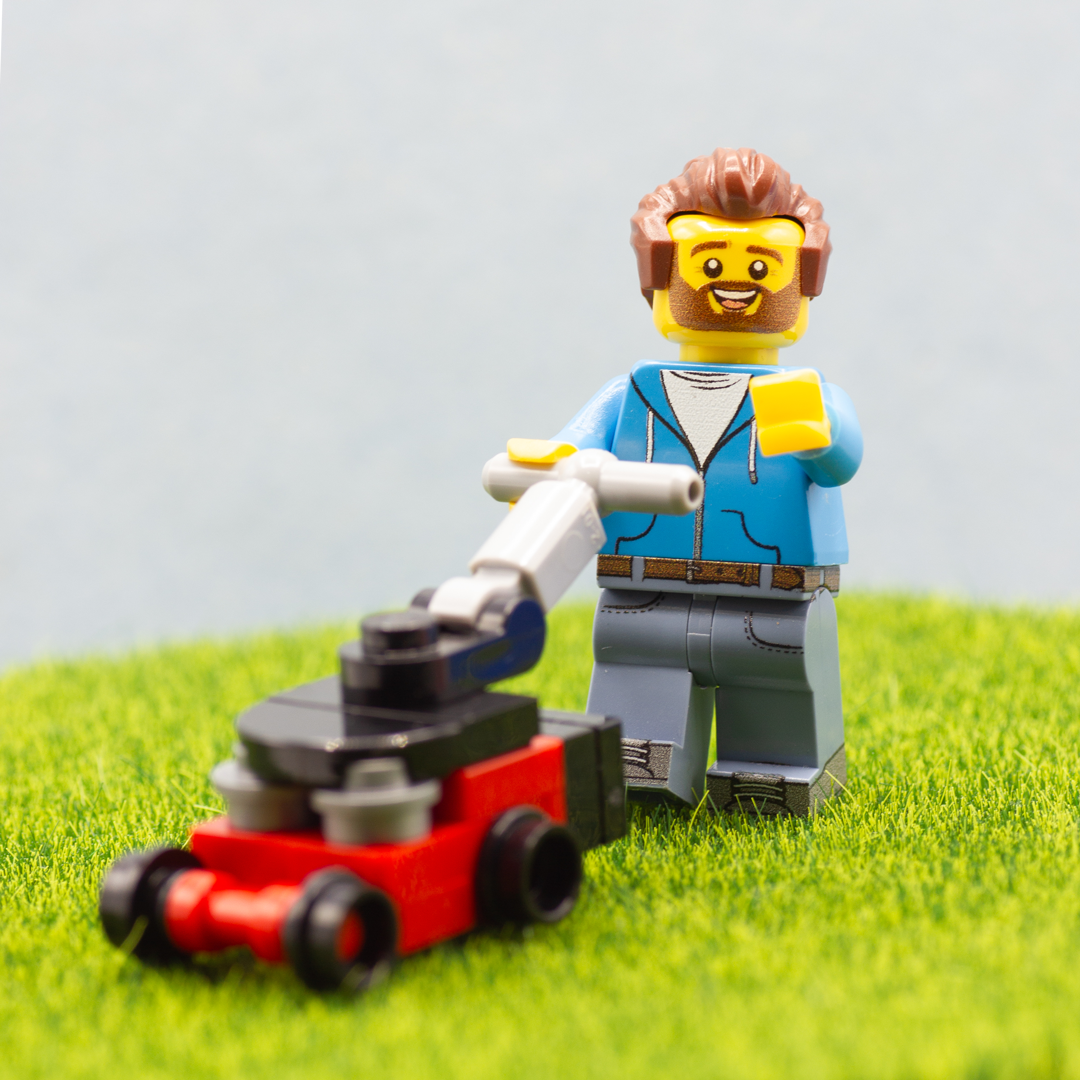 Happy custom minifigure man pushing a red, black and grey LEGO lawnmower over artificial grass