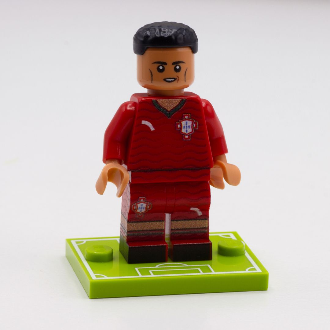 A custom-built toy minifigure of a soccer player with black hair, wearing a red Portugal jersey and shorts, standing on a green soccer field base against a white background.
