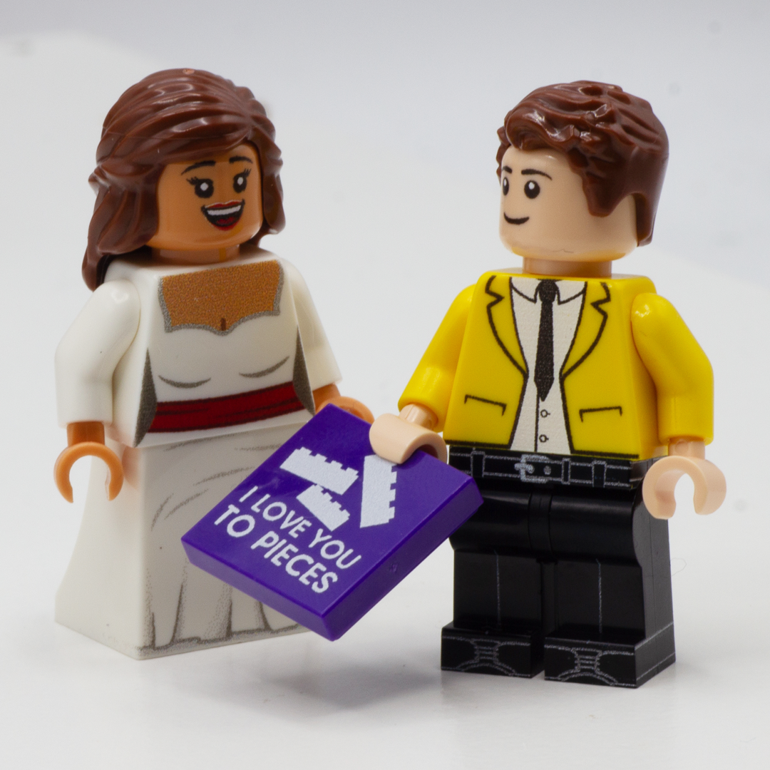 Two custom-built LEGO minifigures holding a purple message tile that reads 'I LOVE YOU TO PIECES' against a clean white background.