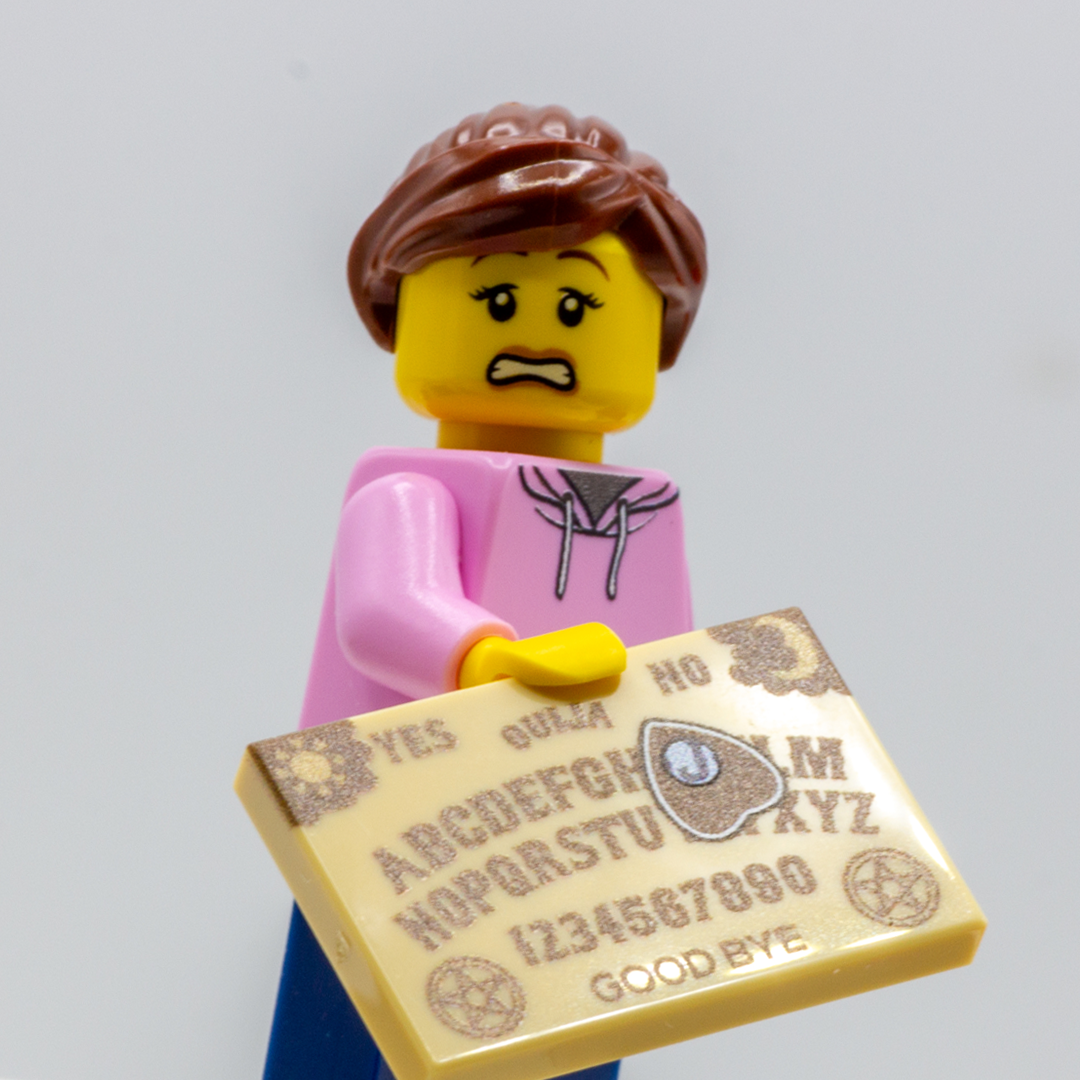 A custom LEGO minifigure with brown hair, a pink top, and blue pants holding a light tan Ouija board against a plain light gray background.