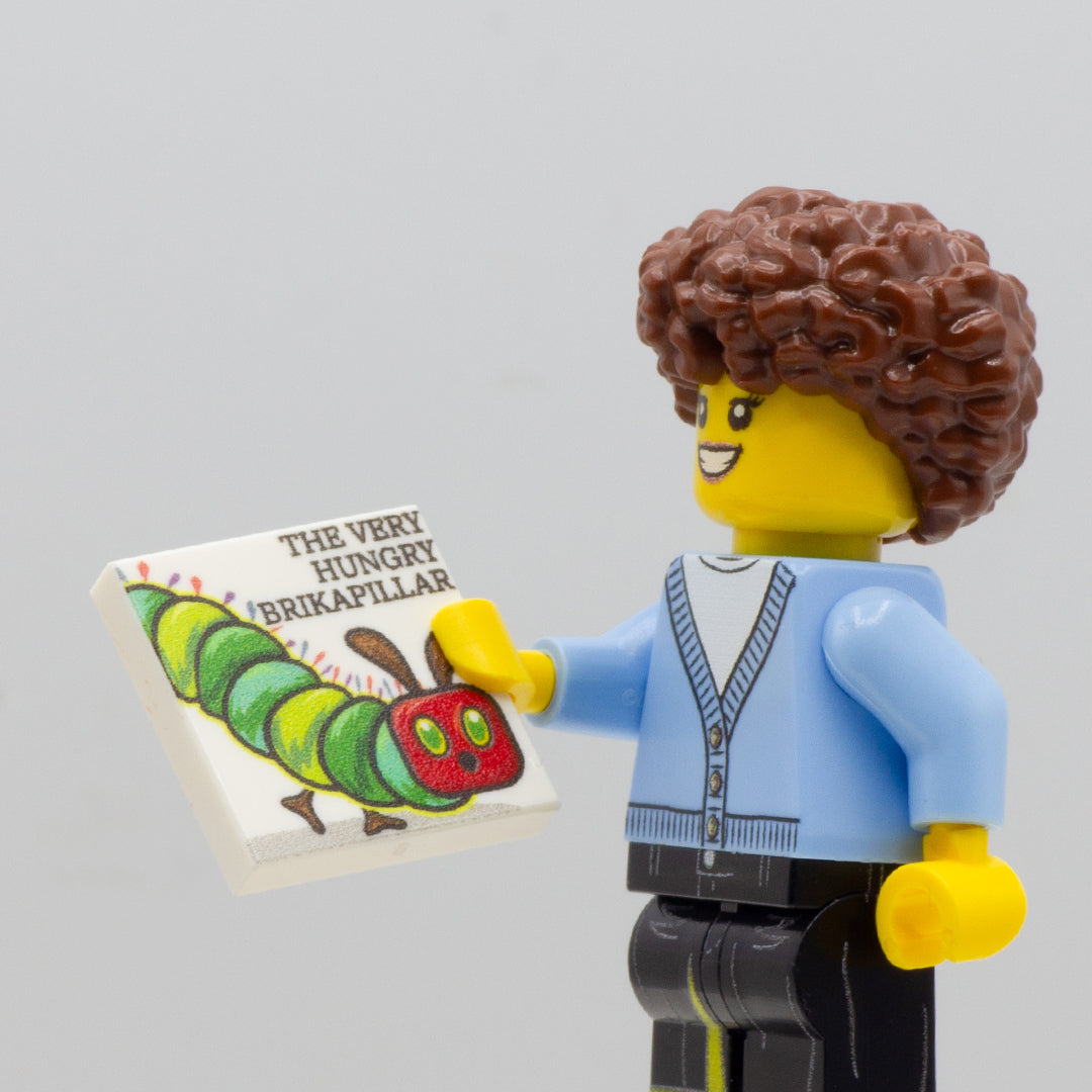 Minifigure holding custom LEGO tile with printed caterpillar on it to resemble a book, The Very Hungry Caterpillar.