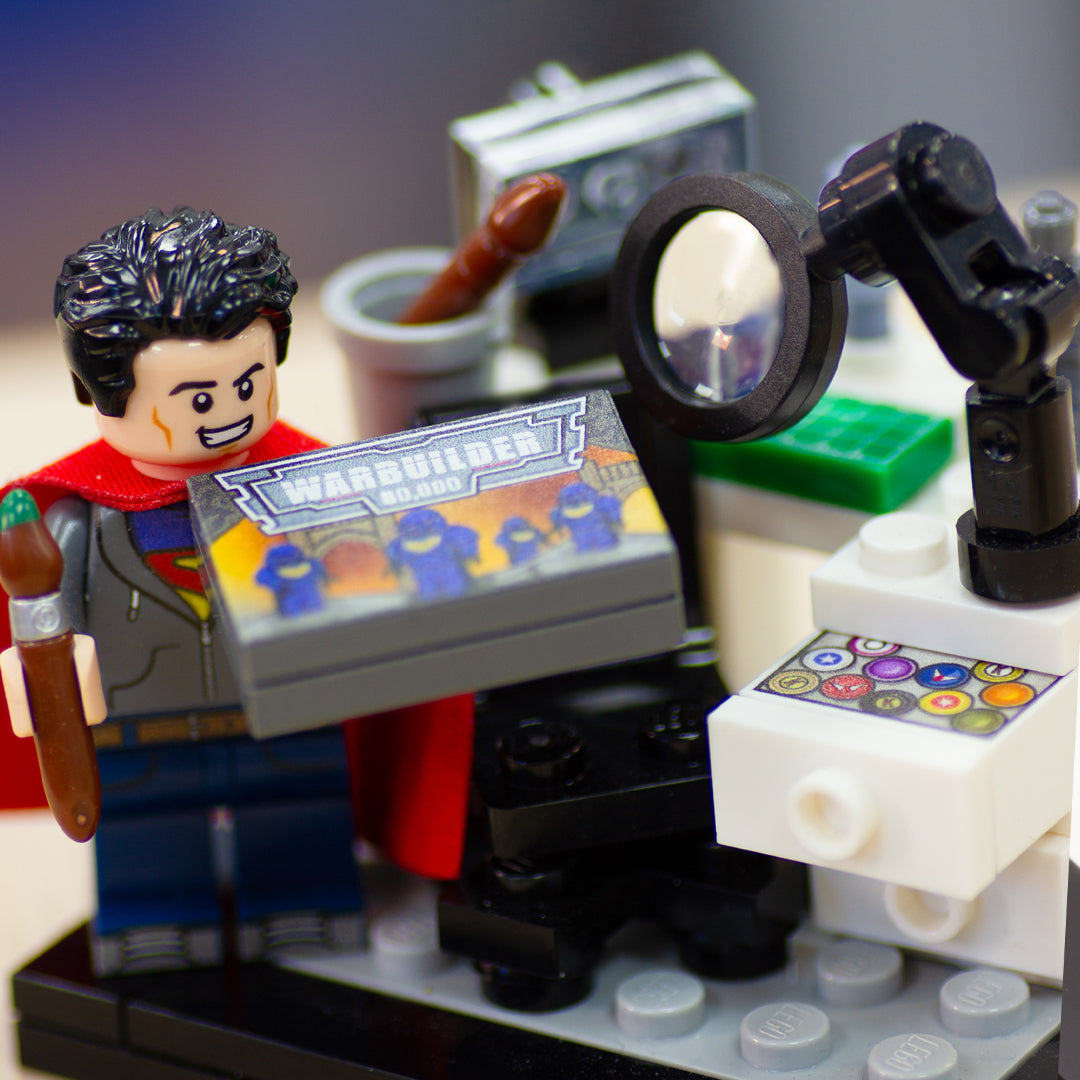 A minifigure inspired by Henry Cavill holding a Warhammer-style box, and standing by a LEGO-style miniature artist's desk set.