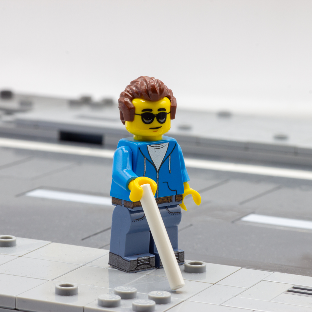 A custom LEGO minifigure with brown hair, black sunglasses, a blue jacket, and blue pants, holding a white stick, standing on a gray brick platform representing a road.